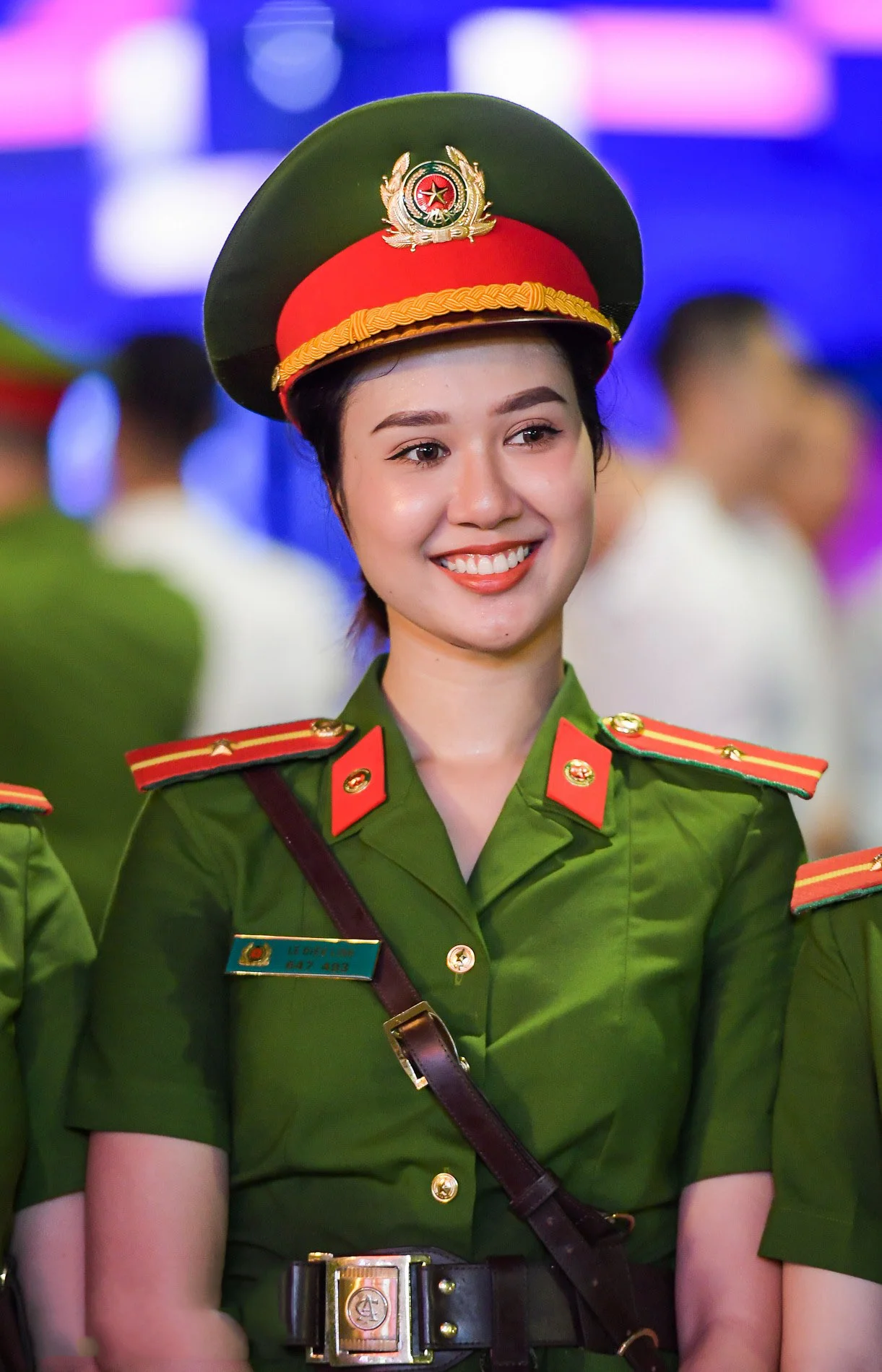 Ảnh công an nữ Việt Nam mặc quân phục xanh đội mũ kêpi và mỉm cười tại một sự kiện trong nhà